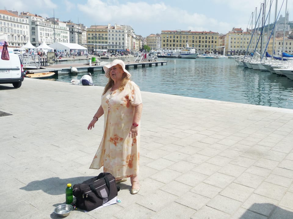 Eine Frau steht an einem Hafen und singt. Im Hintergrund sieht man Boote und Häuser von Marseille. 
