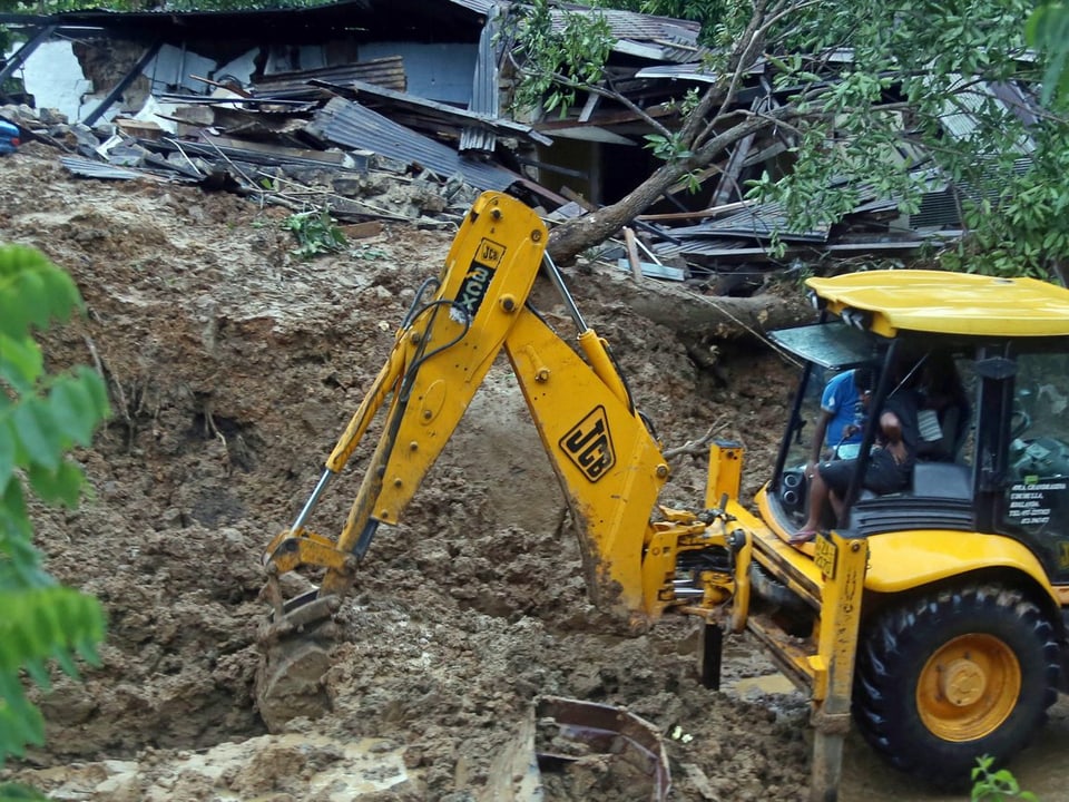 Ein Bagger gräbt im Erdrutschgebiet in Sri Lanka. Dahinter ein zerstörtes Haus.