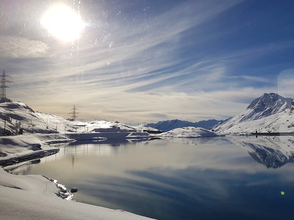 Die verschneite Landschaft auf dem Berninapass mit See und Zugstrecke.