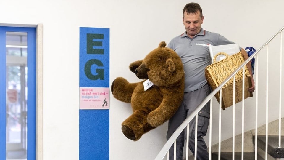 Ein Mann am Zügeln, der einen Korb und einen grossen Teddybären die Treppe hinunter trägt.