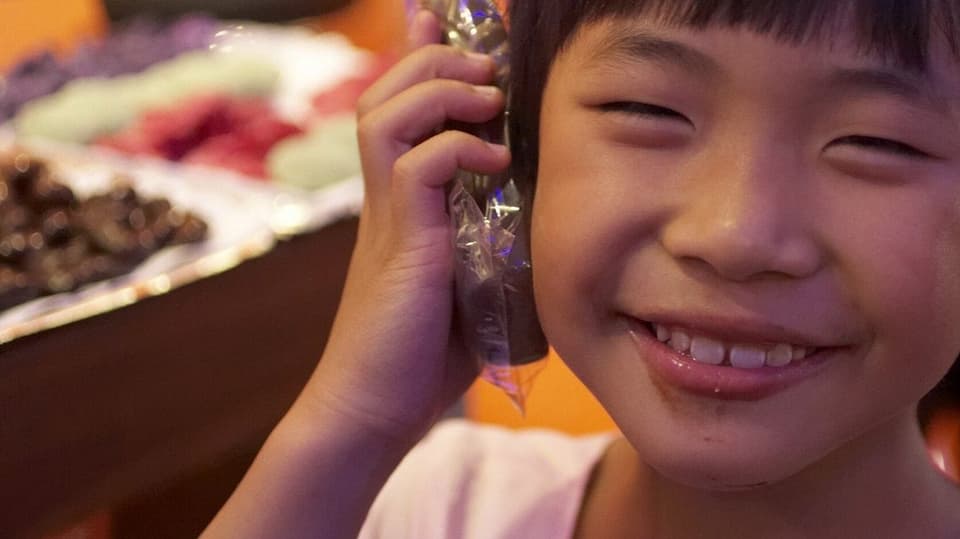 Ein chinesisches Kind hält ein in Zellophan verpacktes Stück Schokolade ans Ohr und lacht.