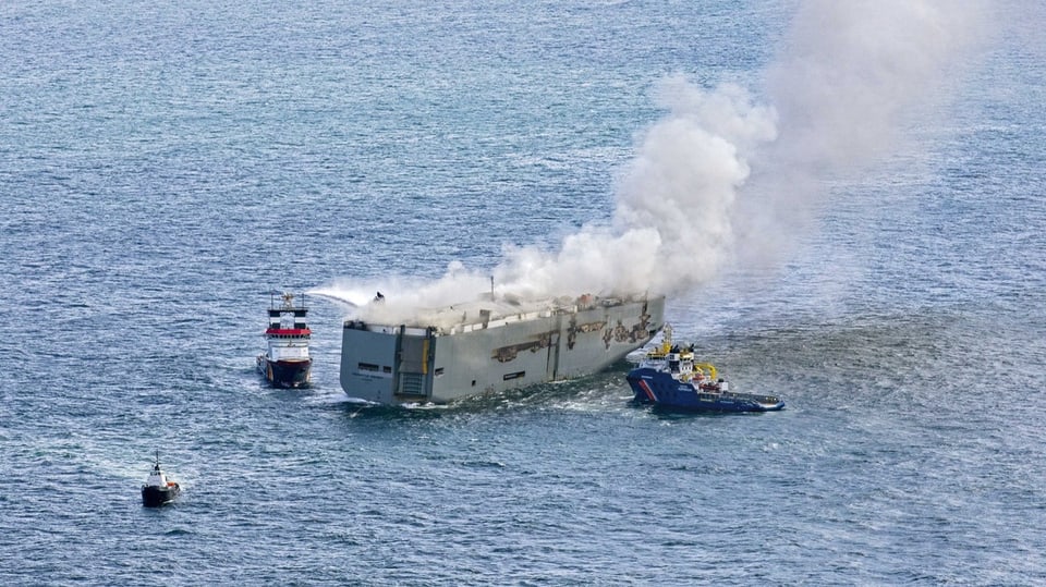 Ein Schiff mit einer Rauchwolche. Drei kleiner Schiffe rundherum, eines versucht den Brand zu löschen.