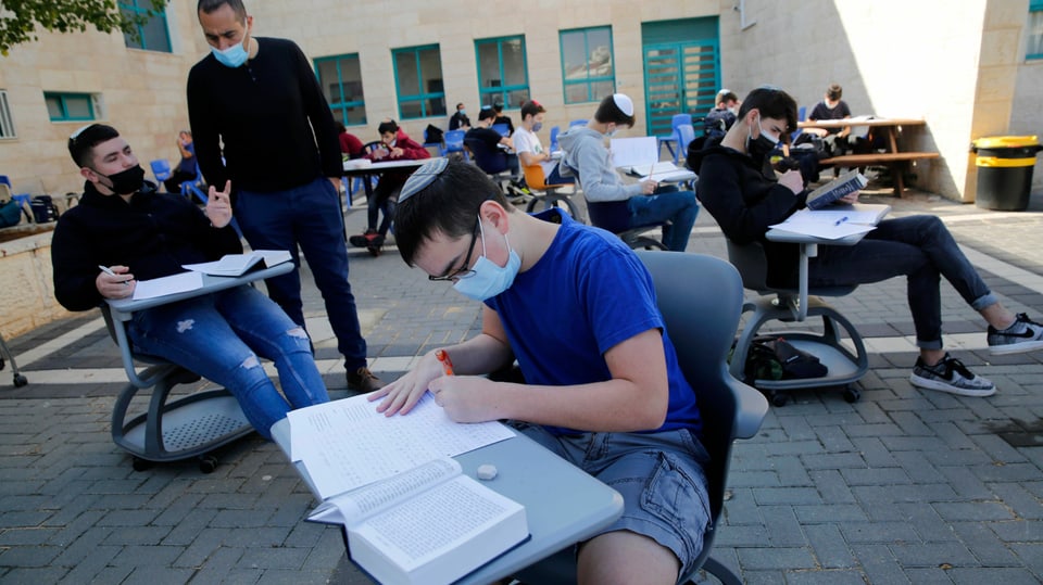 Schüler mit Kippa sitzen auf Bürostühlen auf einem Platz. Sie haben Tischchen vor sich, auf denen Bücher liegen.