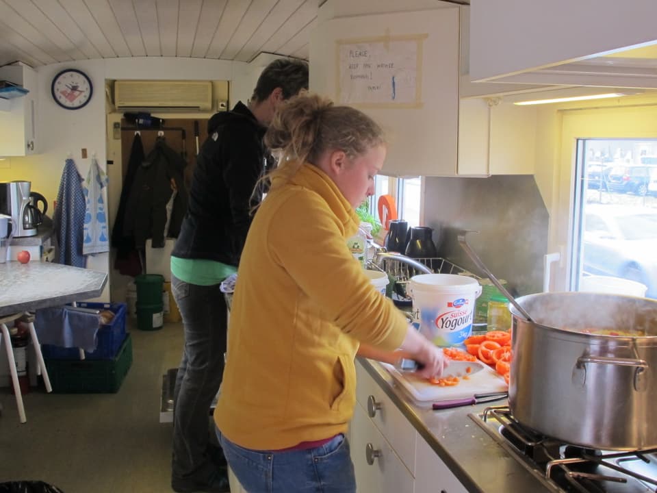 Zwei Personen sind in einem Wohnwagen mit einer kleinen Küche am Kochen mit grossen Töpfen.