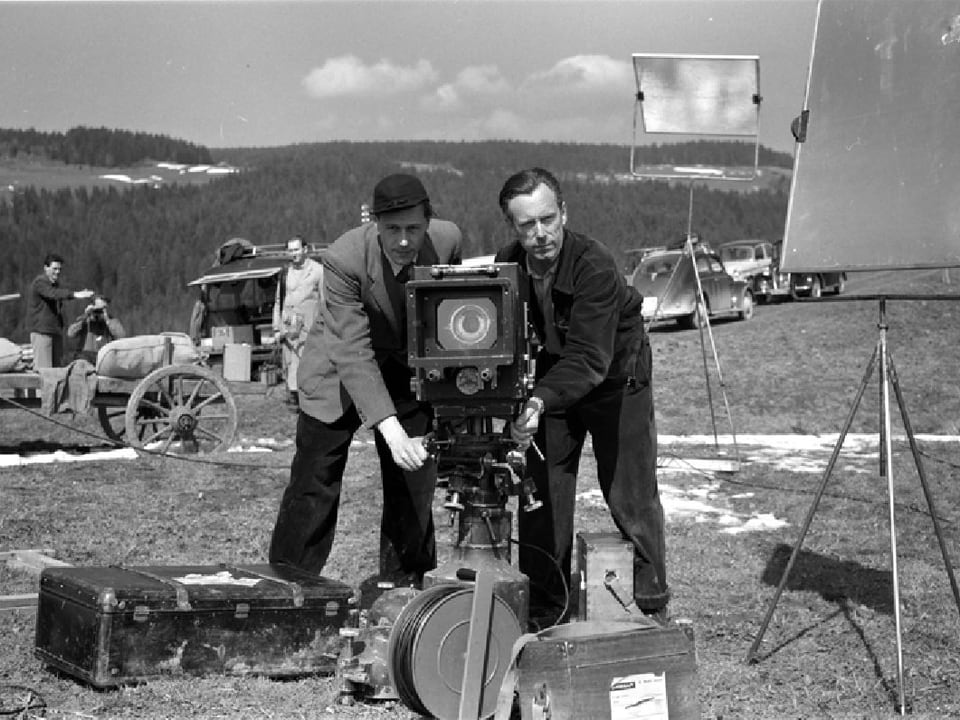 Der Kameramann und sein Assistent beim Ausrichten der Kamera auf einem Feld. Hinter ihnen stehen zwei Lichtreflektoren.