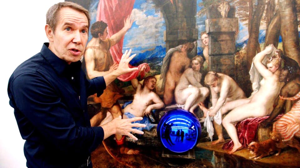 Jeff Koons steht vor einem Gemälde. Er gestikuliert.