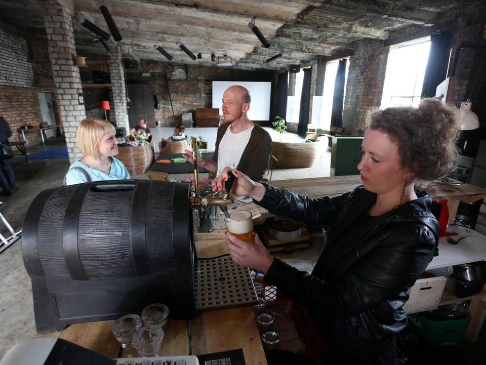 Bar in einer alten Fabrikhalle. Eine Frau zapft Bier aus einem Holzfass.