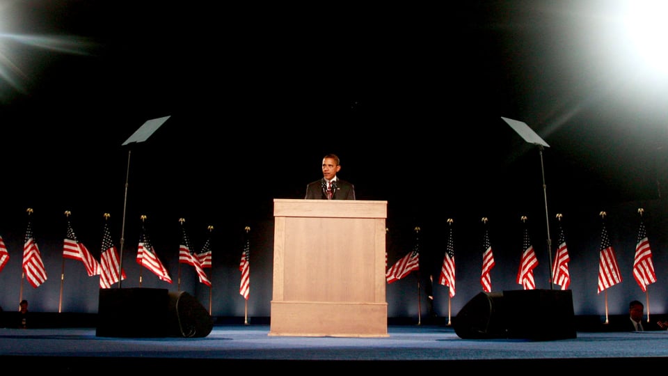 Barack Obama steht an einem Rednerpult, dahinter Amerika-Flaggen.