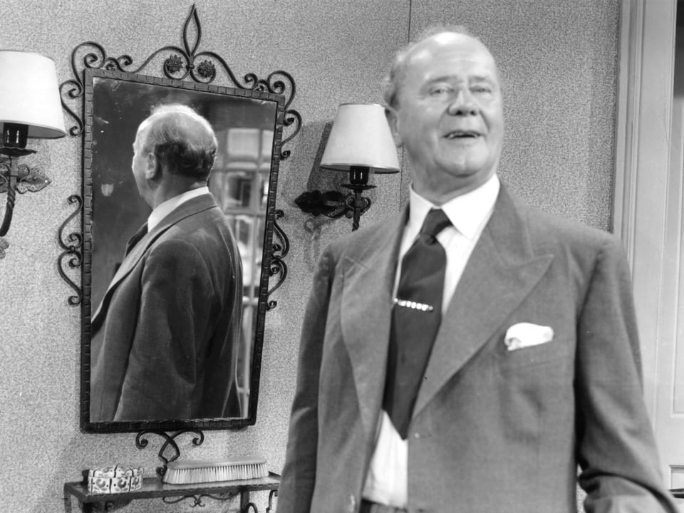 Ein Mann steht mit dem Rücken zu einem Spiegeltisch und lächelt in die Kamera.