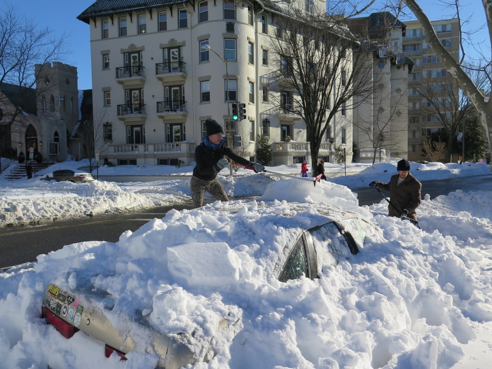 Zwei Menschen schaufeln ein mit Schnee beladenes Auto frei 