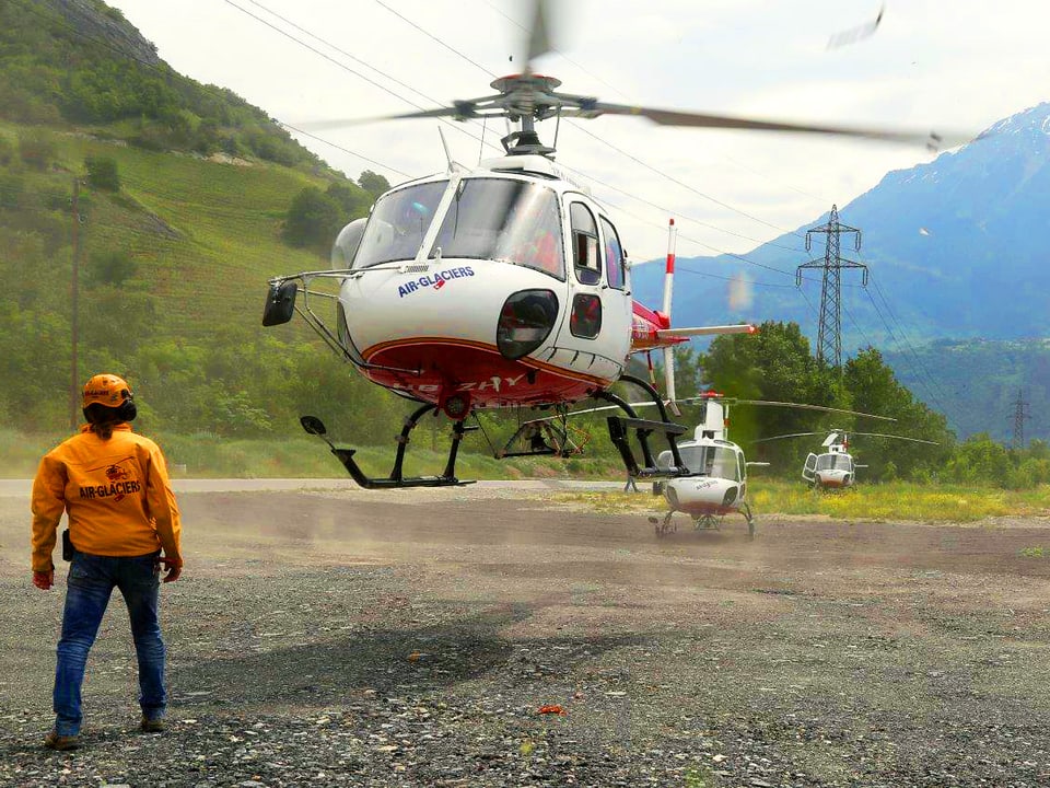 Ein Helikopter kurz vor dem Start auf einem Kiesfeld, daneben steht ein Mann. 