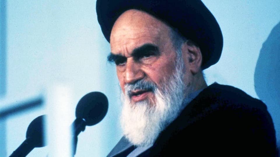 Ayatollah Chomeini mit Turban und Bart vor einem Mikrofon.