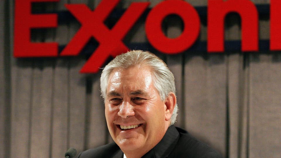 Rex Tillerson lacht in die Kamera vor dem roten Logo von ExxonMobil
