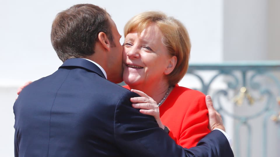 Macron begrüsst Merkel mit Wangenküsschen, Merkel lächelt.