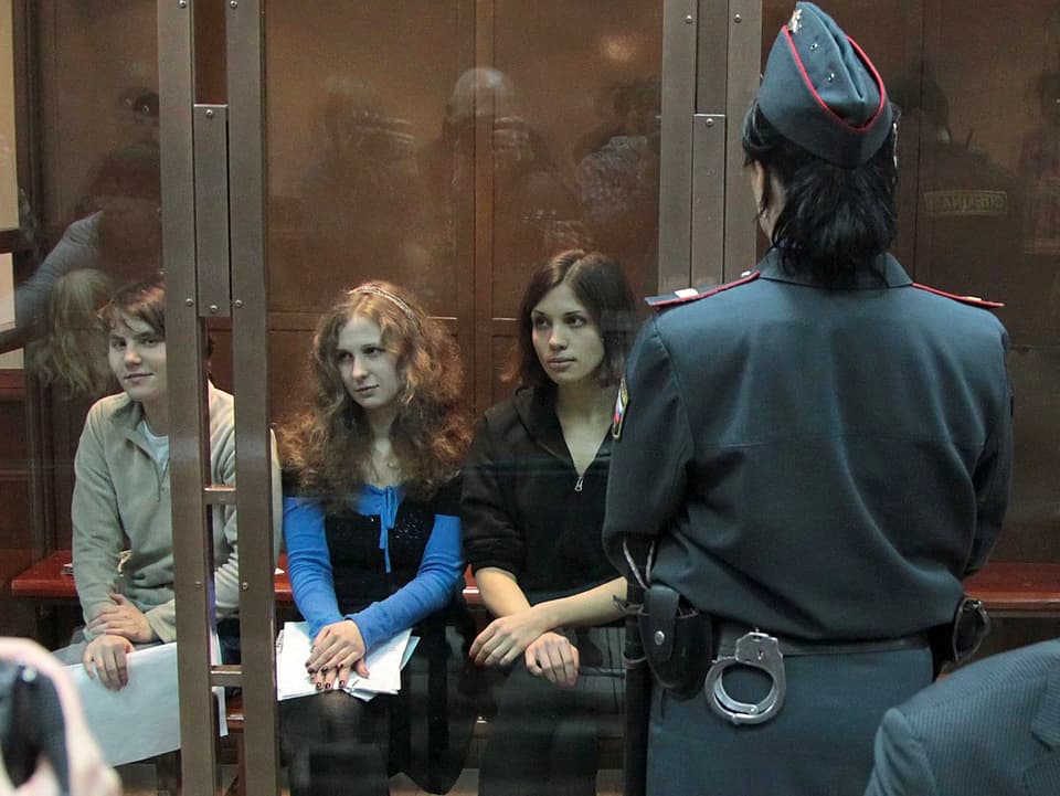 Drei Frauen hinter einer Glaswand sitzend. Davor steht eine Polizistin