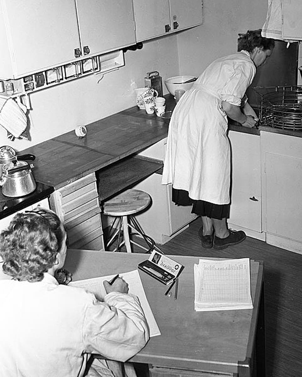 Eine Frau in der Küche, jemand sitzt daneben am Tisch und schreibt auf ein Papier.