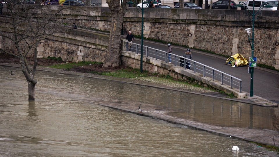 Überfluteter Flussuferweg mit Personen und Bäumen neben einer Mauer