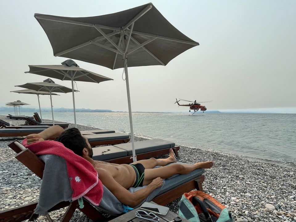 Ein Mann sonnt sich auf einem Liegestuhl am Strand, während im Hintergrund ein Helikopter Wasser auftankt.