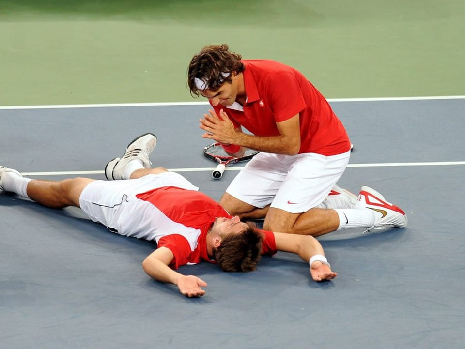 Roger Federer kauert über Stan Wawrinka, der auf dem Court liegt.