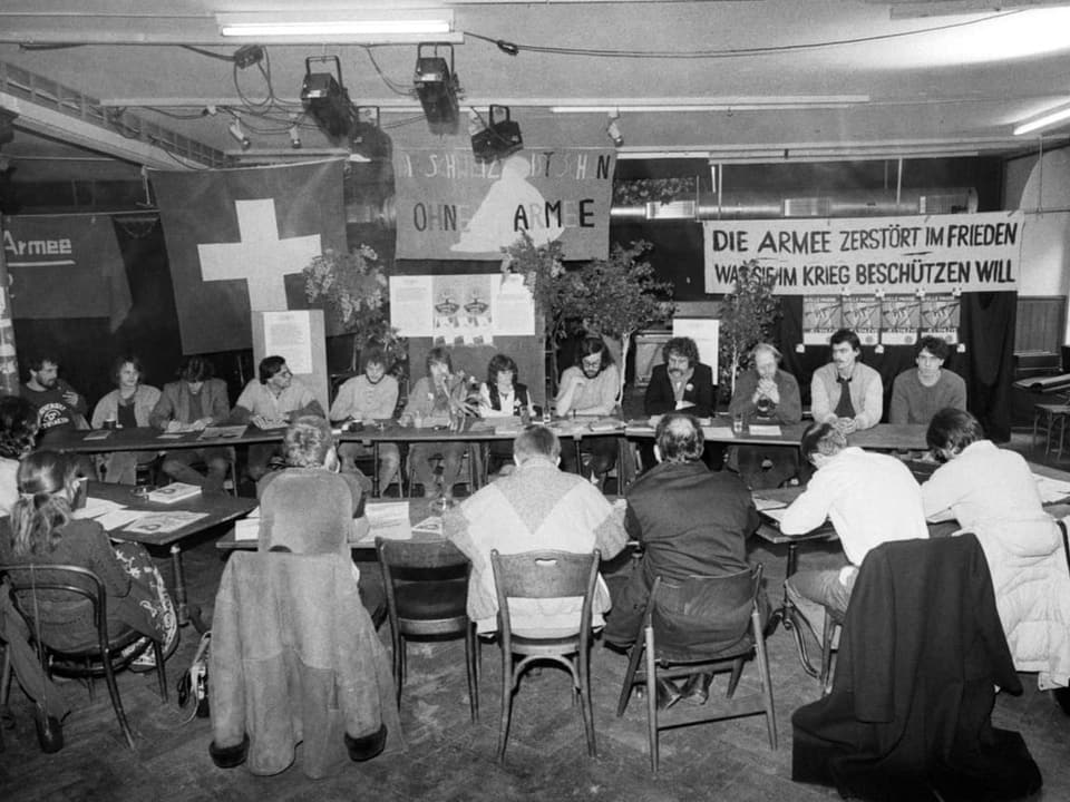 Gruppe von Menschen bei einer Diskussionsrunde in einem Raum mit Plakaten zum Thema Frieden und Armee.