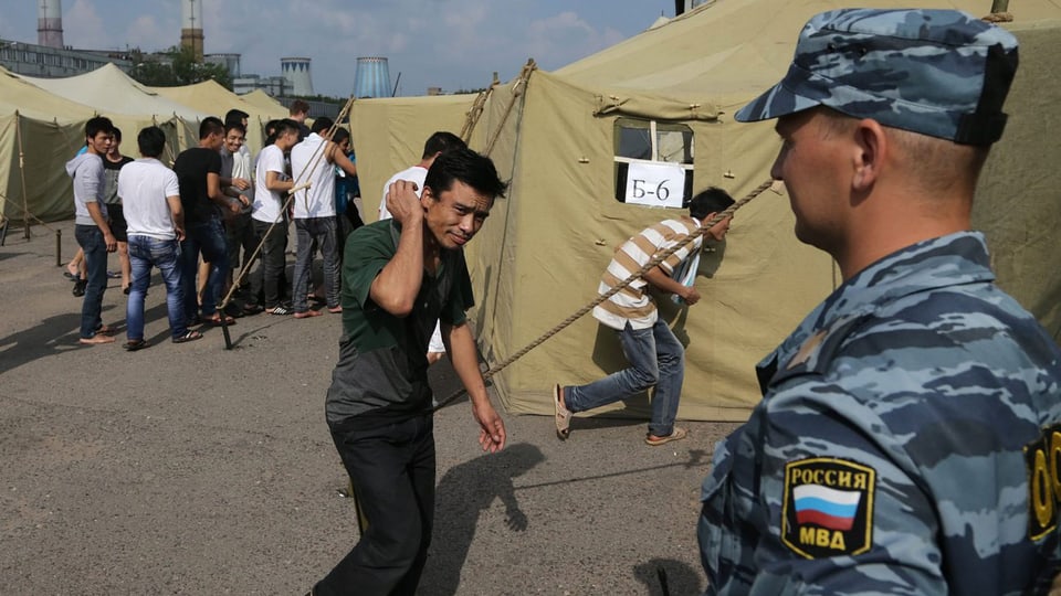 Soldat im Vordergrund. Im Hintergrund asiatische Migranten und Zelte.