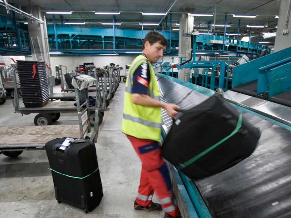 Swissport-Mitarbeiter in gelber Signalweste hebt am Flughafen Zürich einen Koffer auf ein Transportband