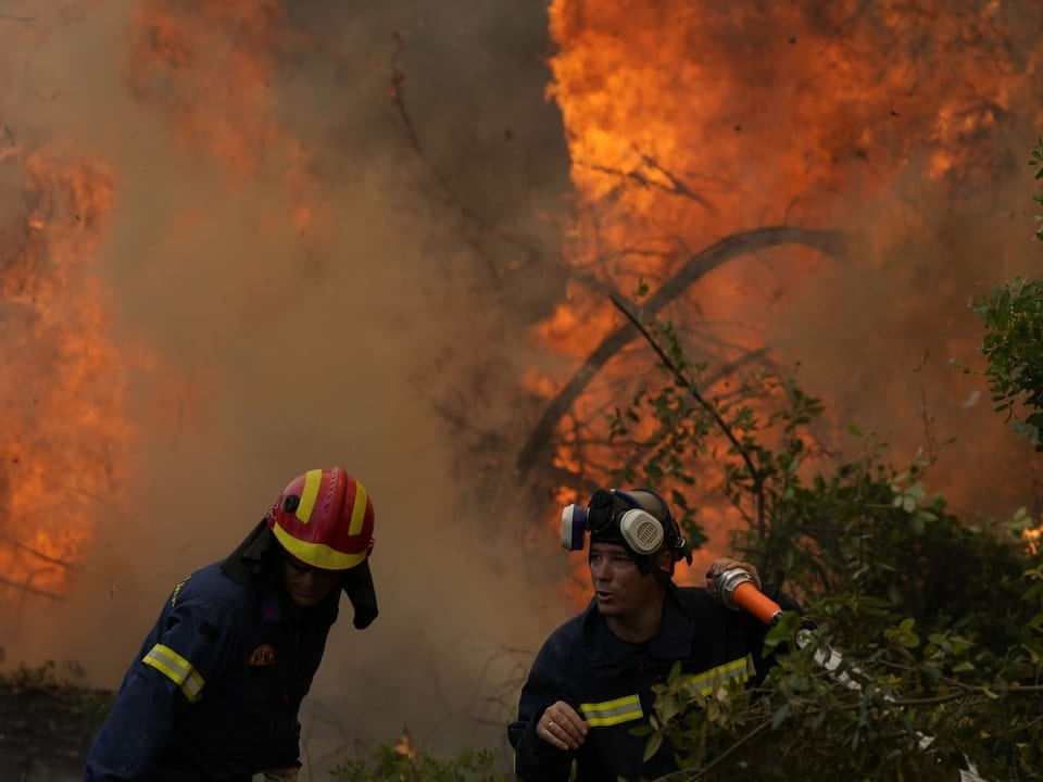 Zwei Feuerwehrmänner stehen mit einem Schlauch in der Nähe eines Feuers.