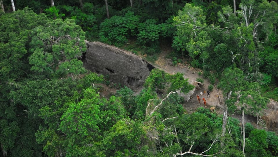 Luftbild mit Indigenen, die in einem Urwald leben. Zu sehen sind einfache Hütten und Menschen.