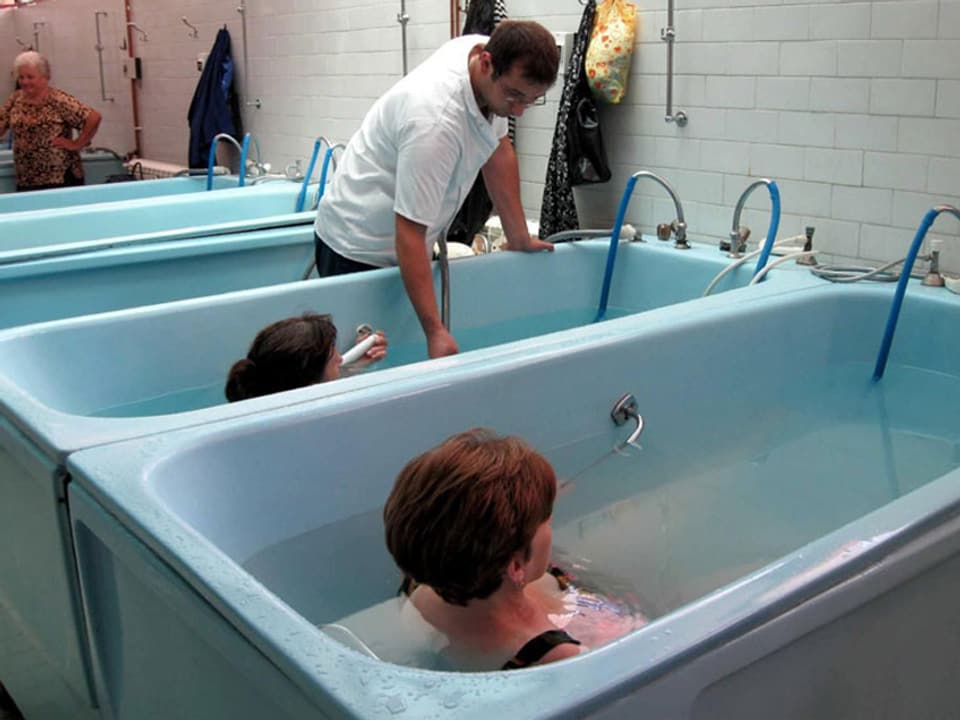Zwei Frauen in grossen Badewannen. Ein Mann in weissem T-Shirt fügt der einen Wanne mehr Wasser zu. 