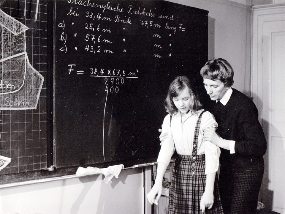 Ein Mädchen steht in einem Schulzimmer vor der Wandtafel. Hiter ihr steht ein Lehrer, der es an beiden Oberarmen festhält.