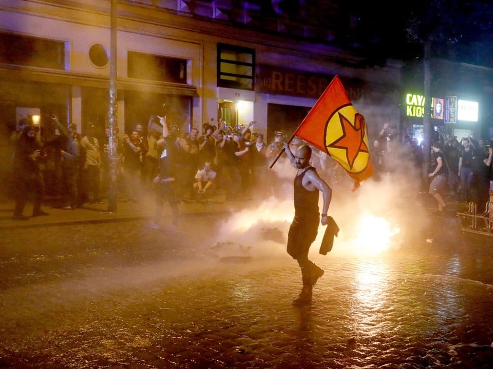 Ein Demonstrant mit einer Fahne, hinter ihm brennt ein Bennsatz.
