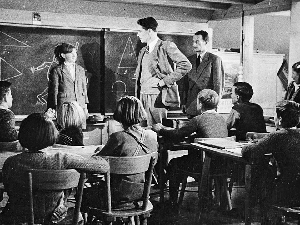 Ein Junge steht vor der Wandtafel vor einer Klasse, neben ihm stehen zwei Männer.