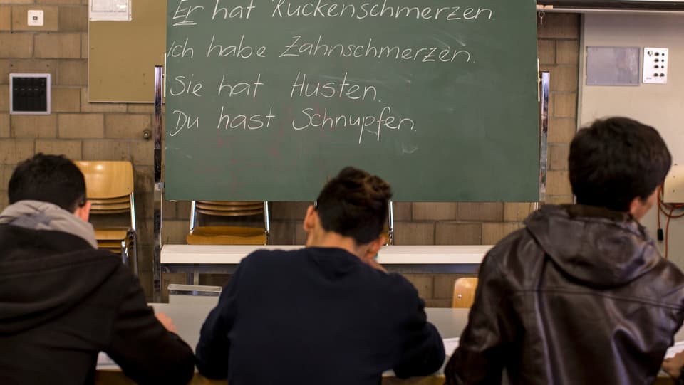 Drei Asylbewerber an einem Tisch von hinten fotografiert, vor ihnen eine Schultafel mit deutschen Sätzen
