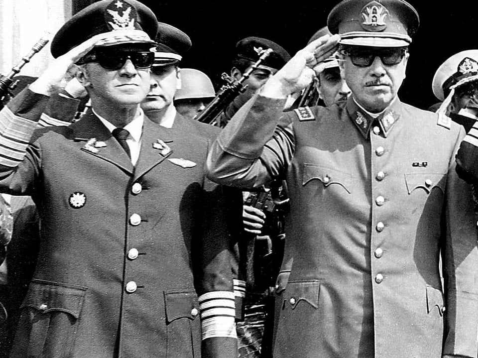 Bild in schwarz/weiss. Augusto Pinochet rechts im Bild und der damalige Kommandant der chilenischen Luftwaffe Gustavo Leigh (links im Bild) salutieren in Uniformen an einer Feier zum Unabhängigkeitstag. 