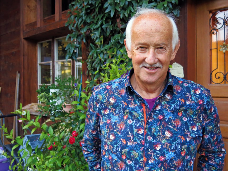 Porträt von Mann in geblümten Hemd vor Holzhaus mit grünen Pflanzen.