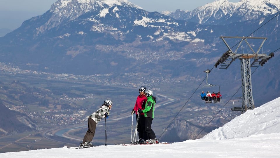 Skifahrer auf der Piste, dahinter das grüne Rheintal in der Ferne.