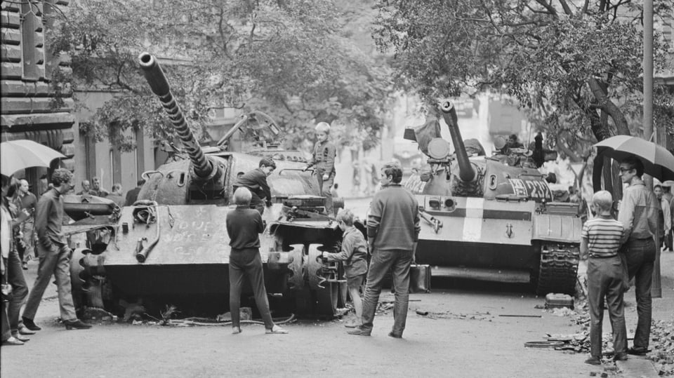 Schwarz-Weiss-Aufnahme zweier Panzer in einer Stadt, die von Passanten gestoppt werden.