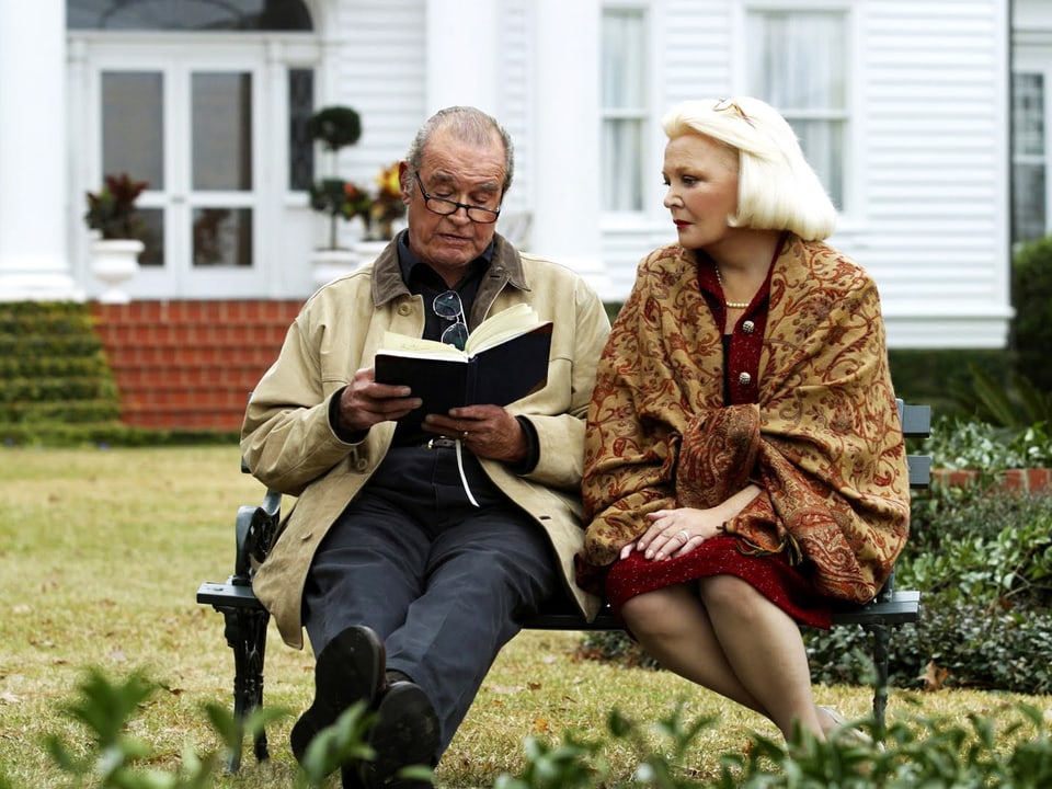 Ein älterer Herr mit einem Buch in der Hand sitzt neben einer älteren Dame auf einer Parkbank.