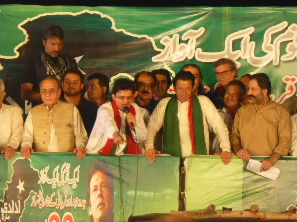 Mehrere Männer auf einer Bühne, ein Mann mit Mikrtofon, neben ihm Imran Kahn mit grünem Schal.