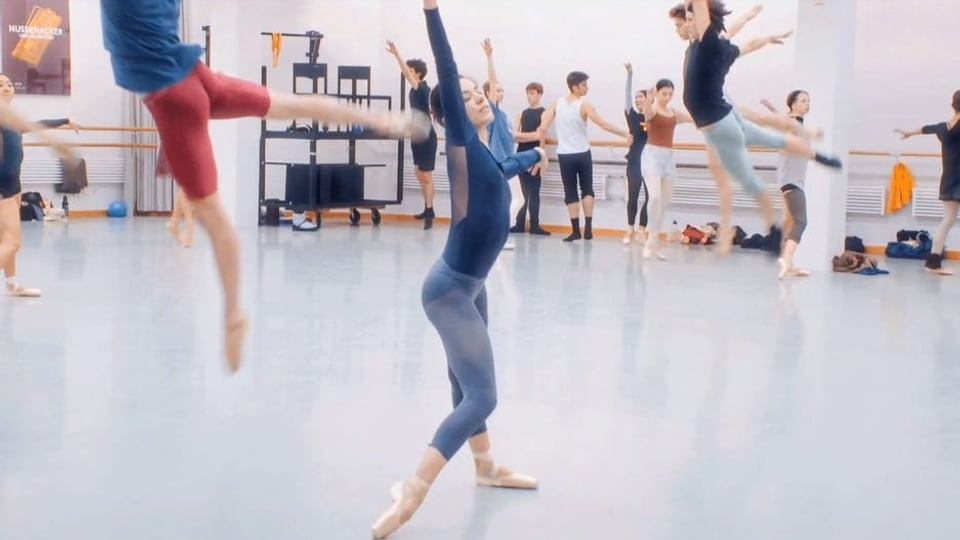 ein Proberaum voller Ballett-Tänzer*innen; im Zentrum tanzt eine Frau.