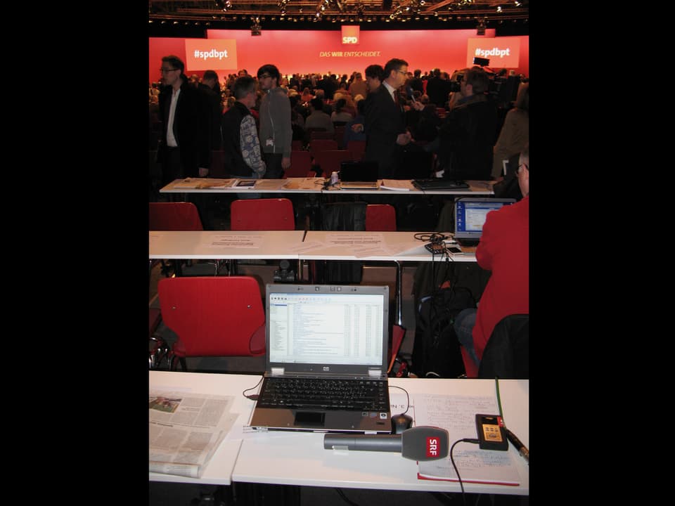 Laptop und Mikrofon auf dem Tisch im Vordergrund, im Hintergrund zwei weitere Tischreihen und viele Leute.