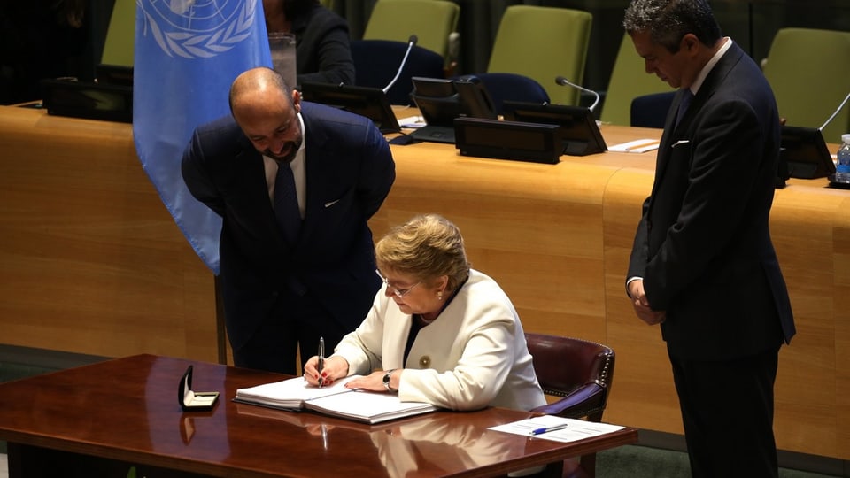 Sie sitzt im weissen Jackett an einem Tisch. Neben ihr stehen zwei Männer, dahinter die UNO-Flagge.