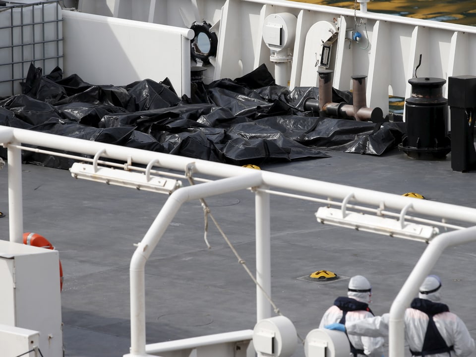 Zwei Beamte in Schutzkleidung sehen aus der Distanz auf zahlreiche Leichensäcke an Bord eines Schiffes