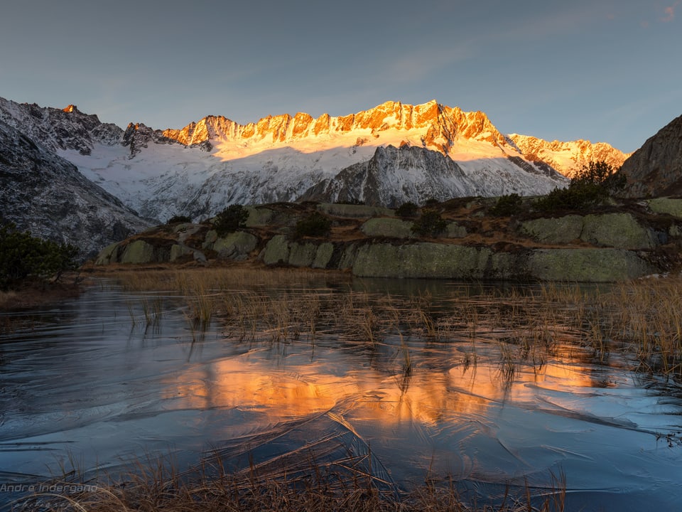 Im Vordergrund ein gefrorener See, dahinter eine Bergkette, die von der Sonne beleuchtet wird.