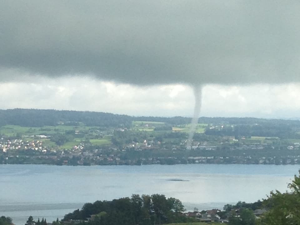 Der Betrachter steht auf einer Anhöhe über dem Zürichsee. Ein Rüssel erstreckt sich von der Wolke bis auf die Seeoberfläche.