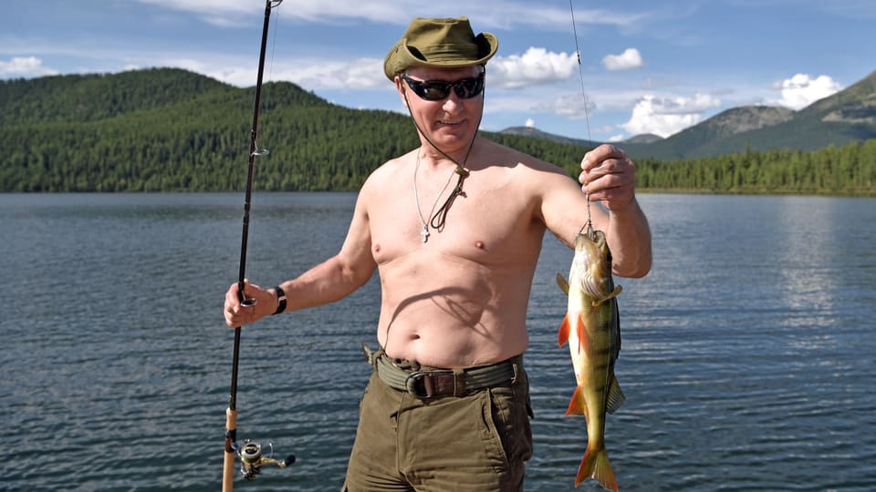 Putin mit Hut und Sonnenbrille hält eine Fischerrute und einen an der Angel hängenden grossen Barsch in den Händen.