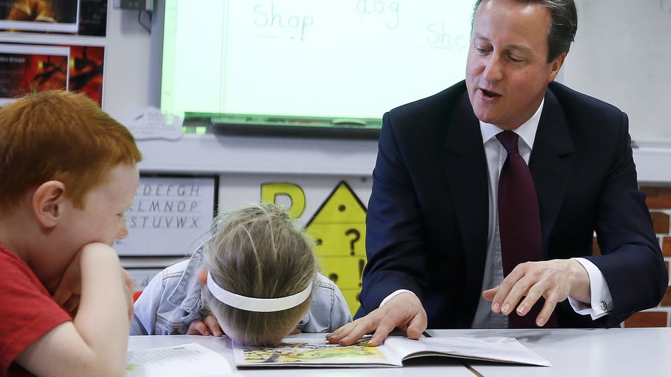 Englands Premier David Cameron sitzt an einem Schultisch, zusammen mit einem kleinen Jungen und einem Mädchen. Das Mädchen hat seinen Kopf mit dem Gesicht nach unten auf den Tisch gelegt.