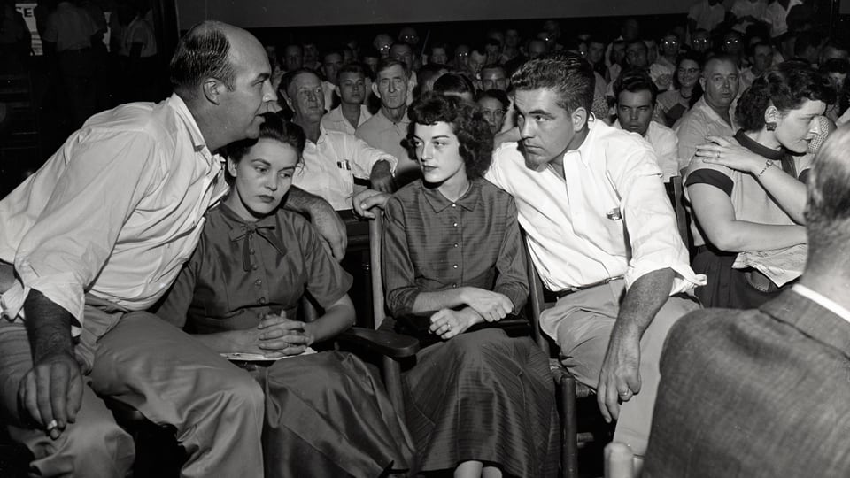 schwarzweiss-Foto von zwei jungen Frauen und zwei Männern auf Stühlen. Einer legt der Frau mit Locken den Arm um