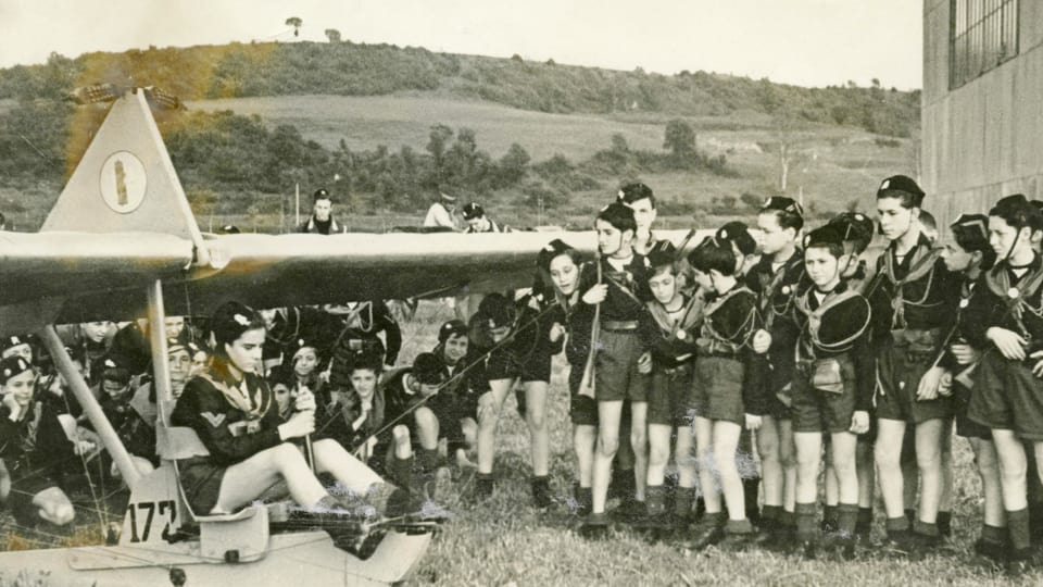 Schwarzweissfoto: Eine Gruppe Kinder in Uniformen steht um einen Gleitflieger herum.