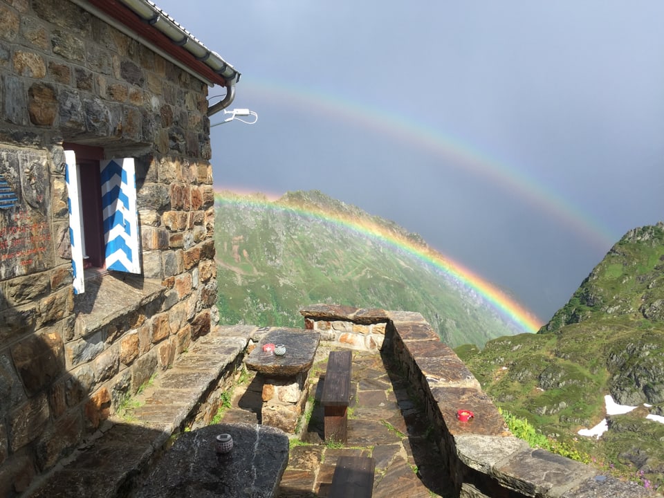 Ein dopelter Regenbogen steht direkt vor der SAC Hütte, die Sonne beleuchtet die Bänke und die Felsen in der näheren Umgebung.